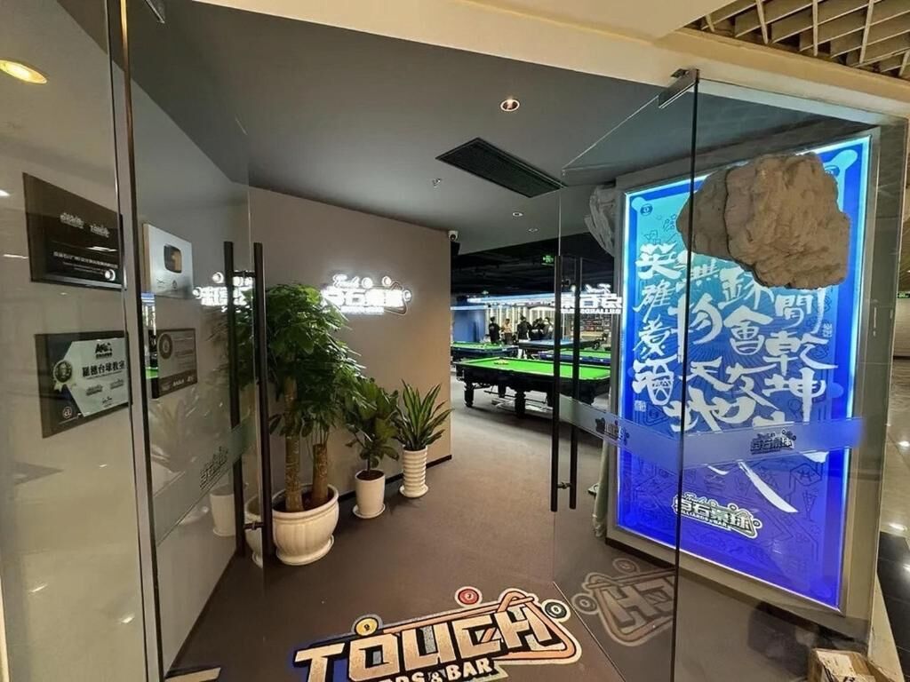 A billiards hall with glass walls, potted plants, and a digital display advertising a game.