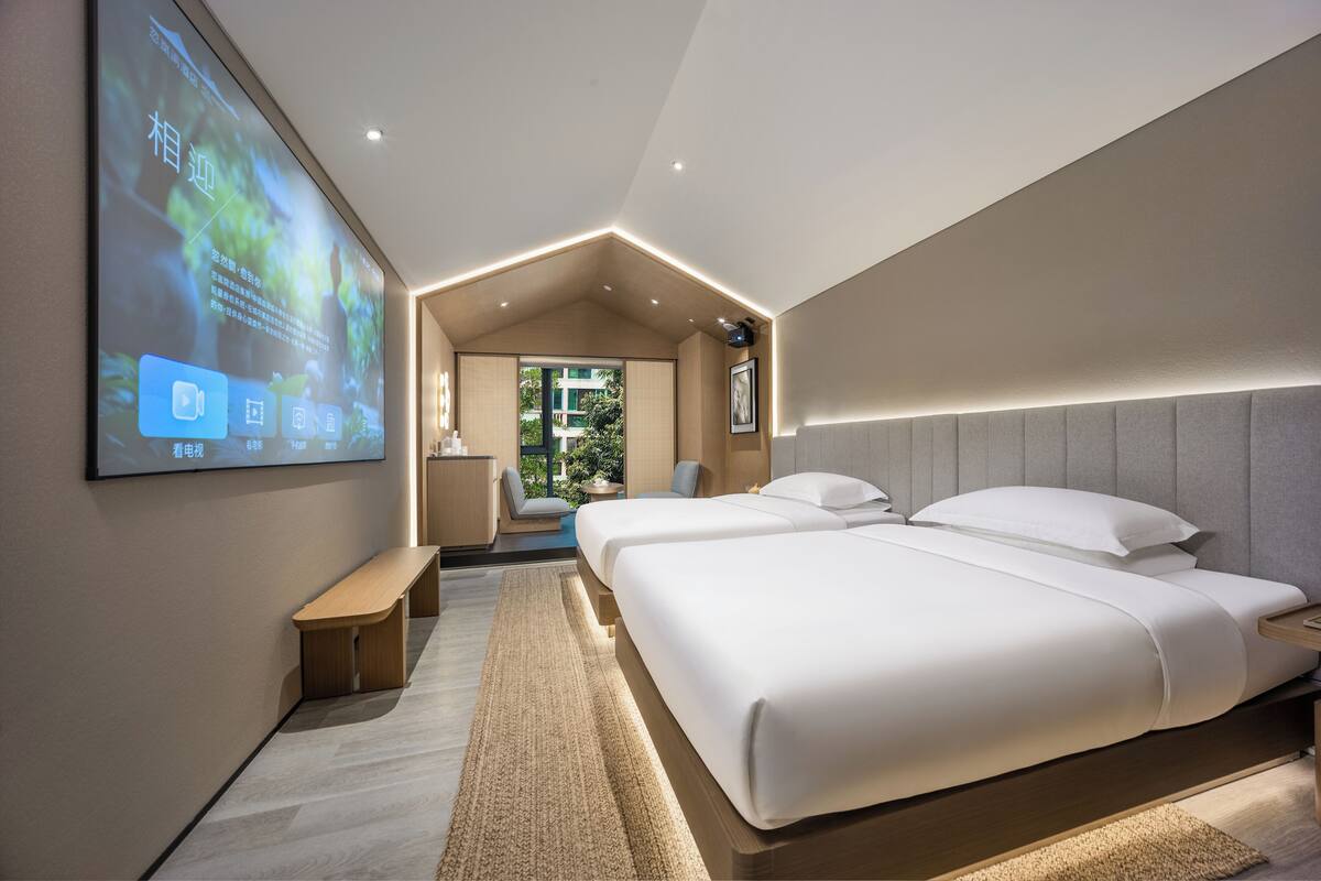 A modern hotel room with a large bed, a desk, and a TV screen displaying content.