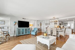 Una sala de estar moderna con un televisor de pantalla plana, una zona de comedor con sillas de mimbre y una cocina con armarios blancos.