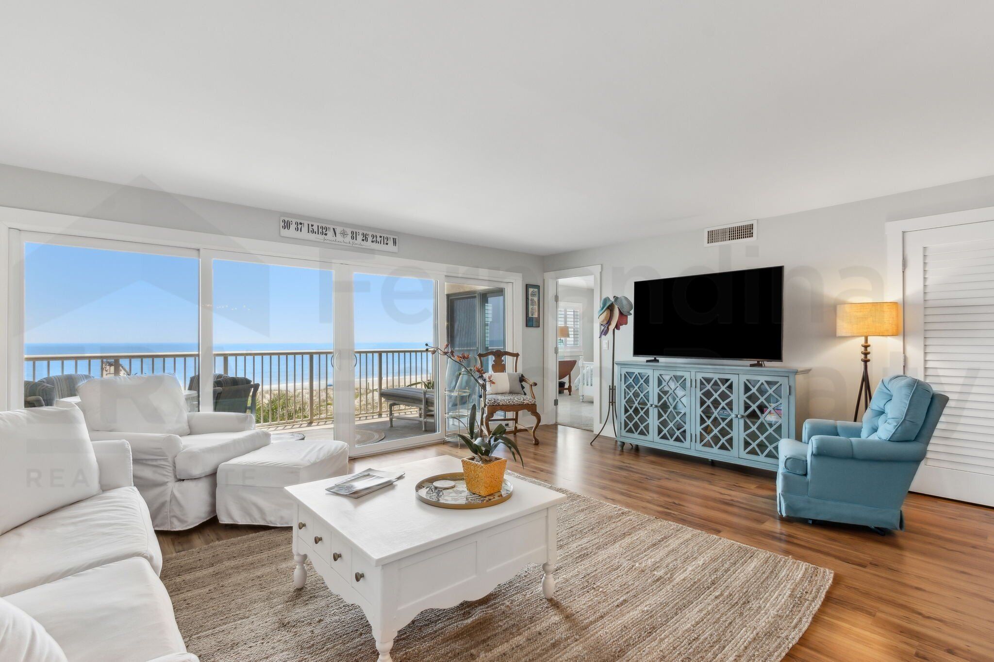 A spacious living room with a white sofa, a coffee table, and a TV. There is a balcony with a view of the ocean.