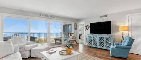 A spacious living room with a white sofa, a coffee table, and a TV. There is a balcony with a view of the ocean.