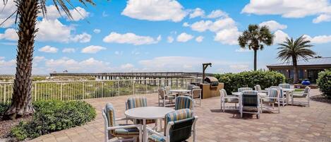 An outdoor patio with round tables and striped chairs, palm trees, and a view of a pier.