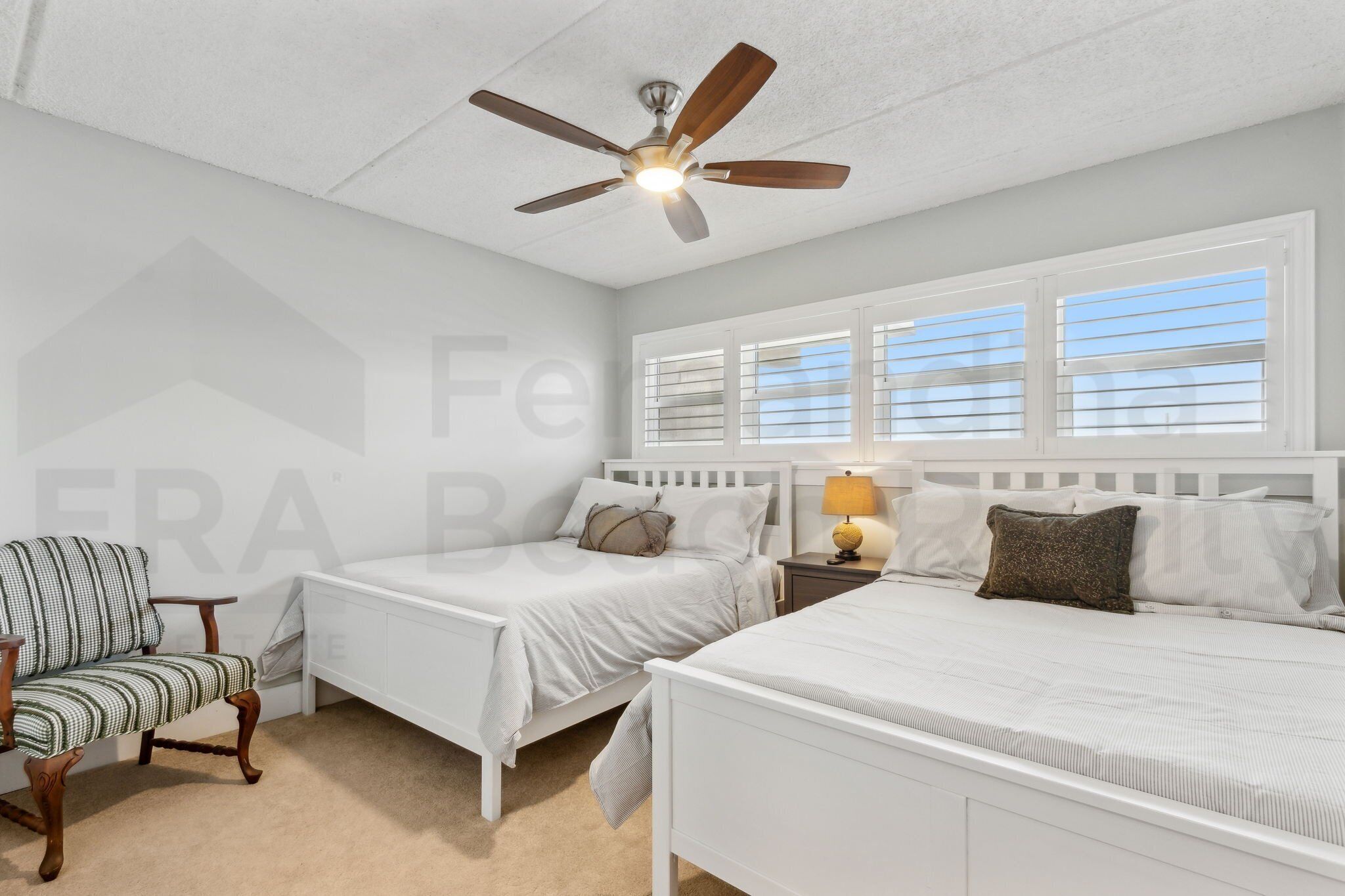 Une chambre avec deux lits simples, un ventilateur de plafond, une table de chevet avec une lampe, une chaise et une fenêtre avec des stores.