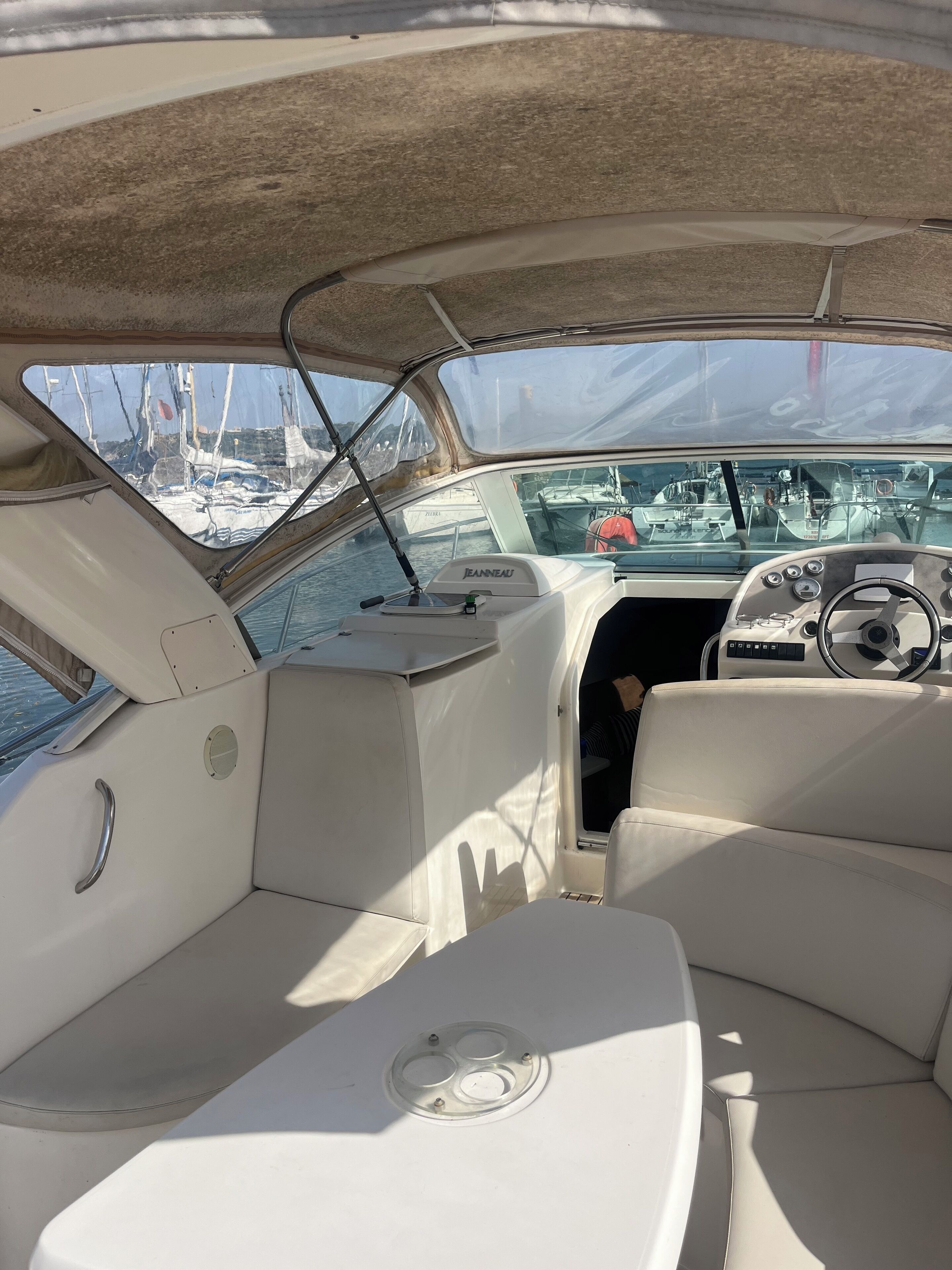 Het interieur van een boot met een stuurwiel, een bedieningspaneel en uitzicht op andere boten in een jachthaven.
