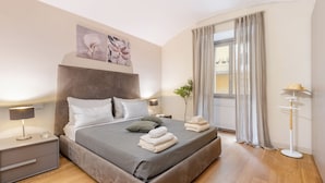 Un dormitorio moderno con una cama grande, mesitas de noche, una lámpara de noche, una planta en maceta y una ventana con cortinas.