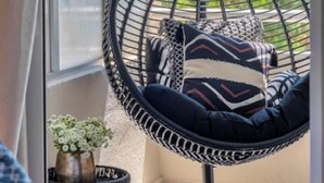 A hanging chair with a cushion, a small table with a vase of flowers, and a view of the sky and buildings in the background. - Voula Urban Escape, Bill & John Villas (Vari - Voula - Vouliagmeni)