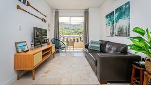 Una sala de estar moderna con un sofá, televisor, mueble de entretenimiento de madera y vistas a un balcón con un paisaje pintoresco.