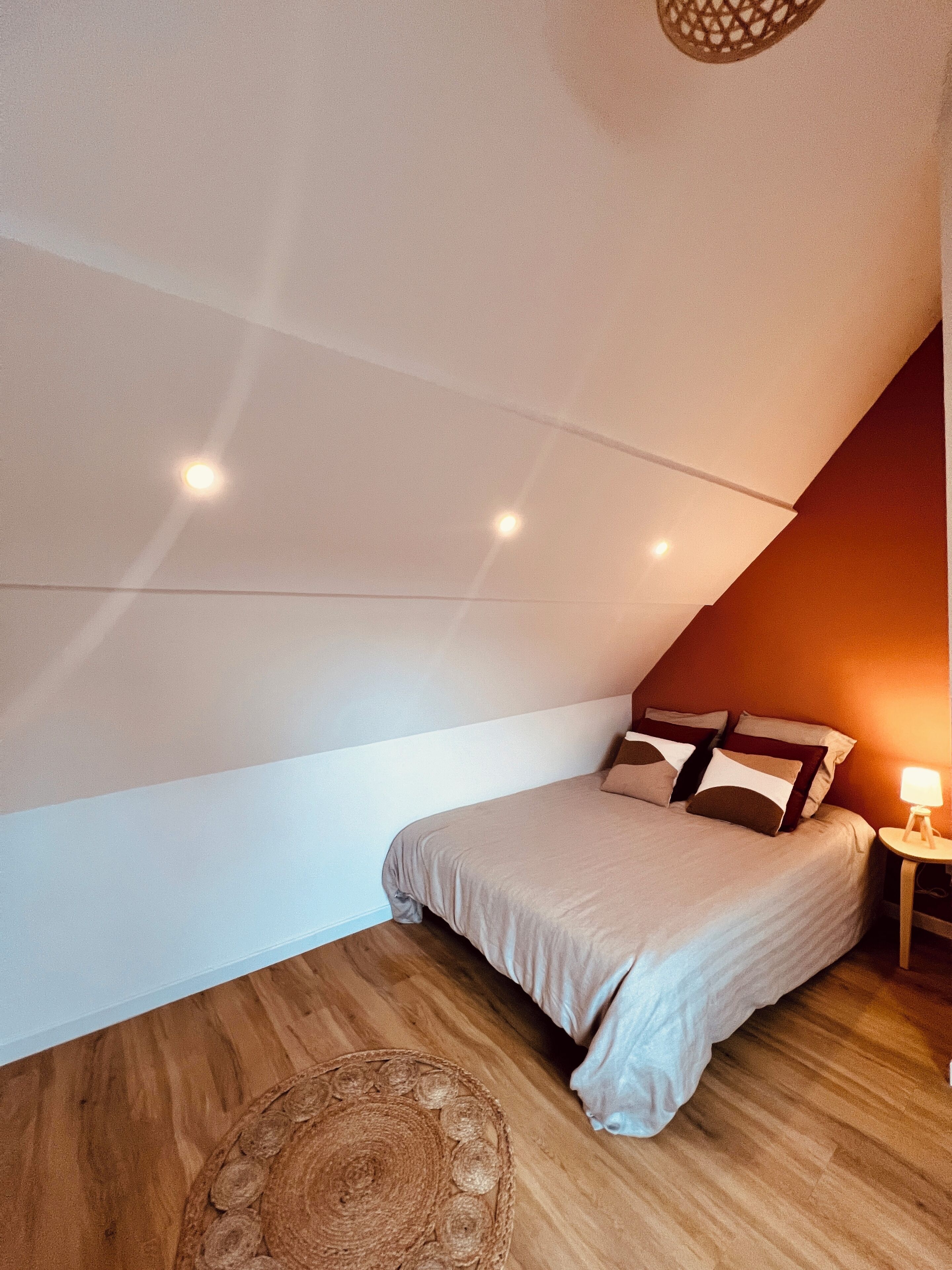 Ein Schlafzimmer mit schrägverdachter Decke, einem Bett mit Kissen, einem Nachttisch mit Lampe und einem gewebten Teppich auf dem Boden.
