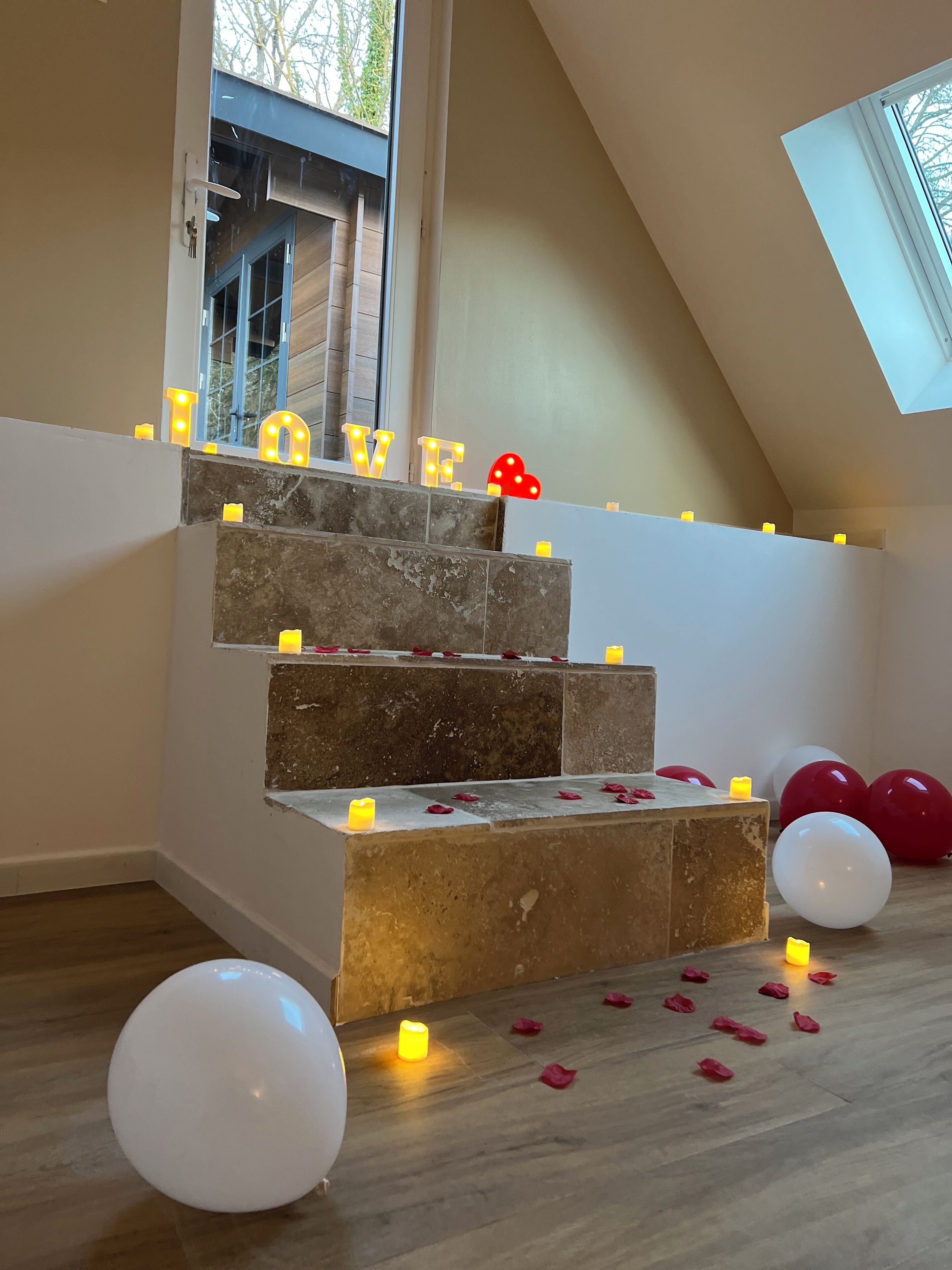 Un escalier décoré de bougies et de ballons, un panneau portant le mot « AMOUR », et une vue sur les arbres à l’extérieur.