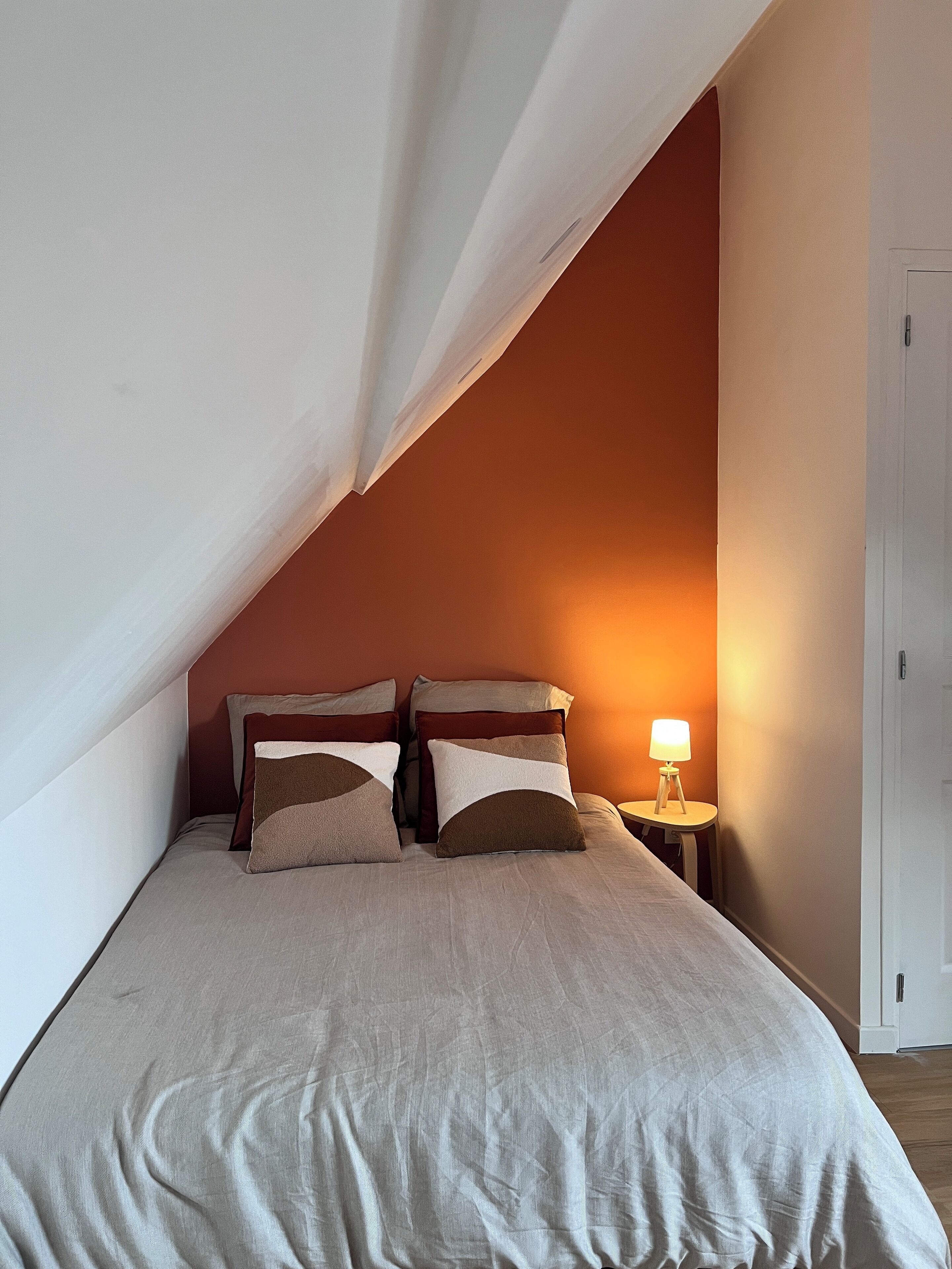 A bedroom with a sloped ceiling, a bed with a beige bedspread, two pillows, and a bedside lamp.