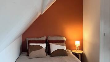 A bedroom with a sloped ceiling, a bed with a beige bedspread, two pillows, and a bedside lamp.