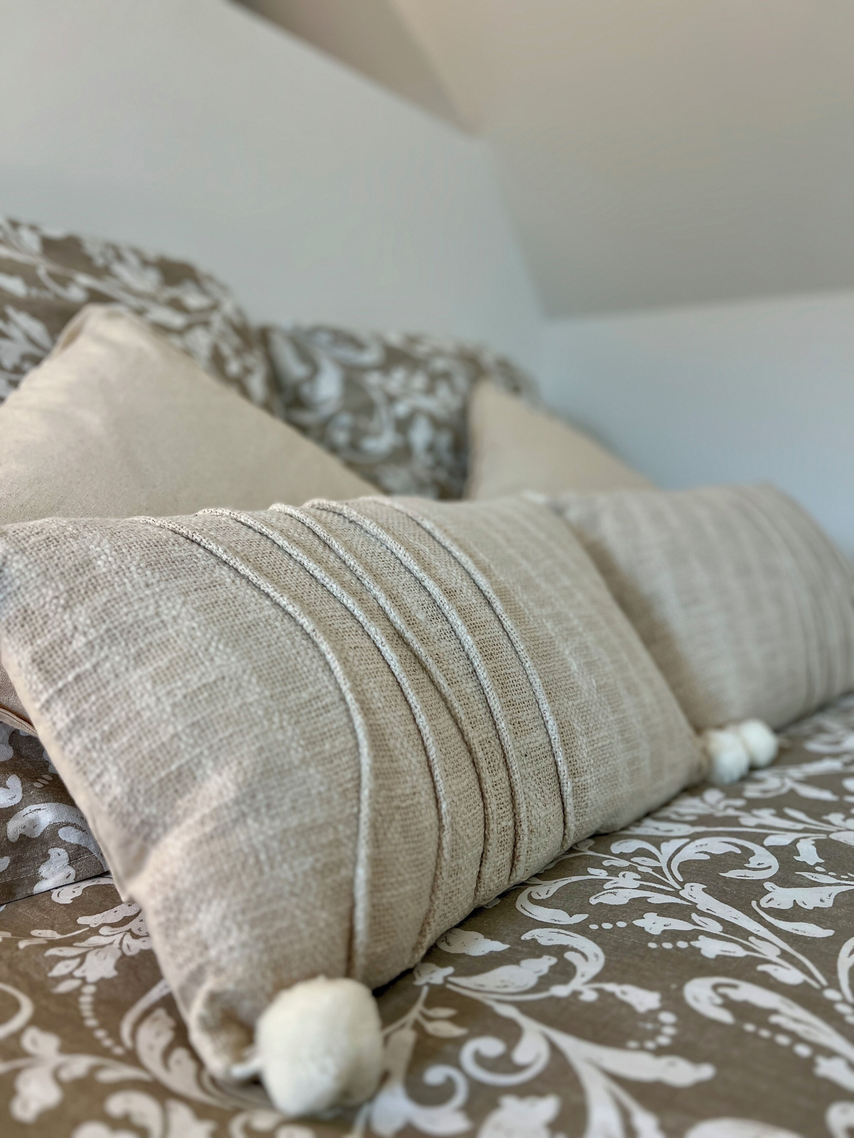 A bed with a patterned bedspread and two beige pillows with a textured fabric and a decorative pom-pom.