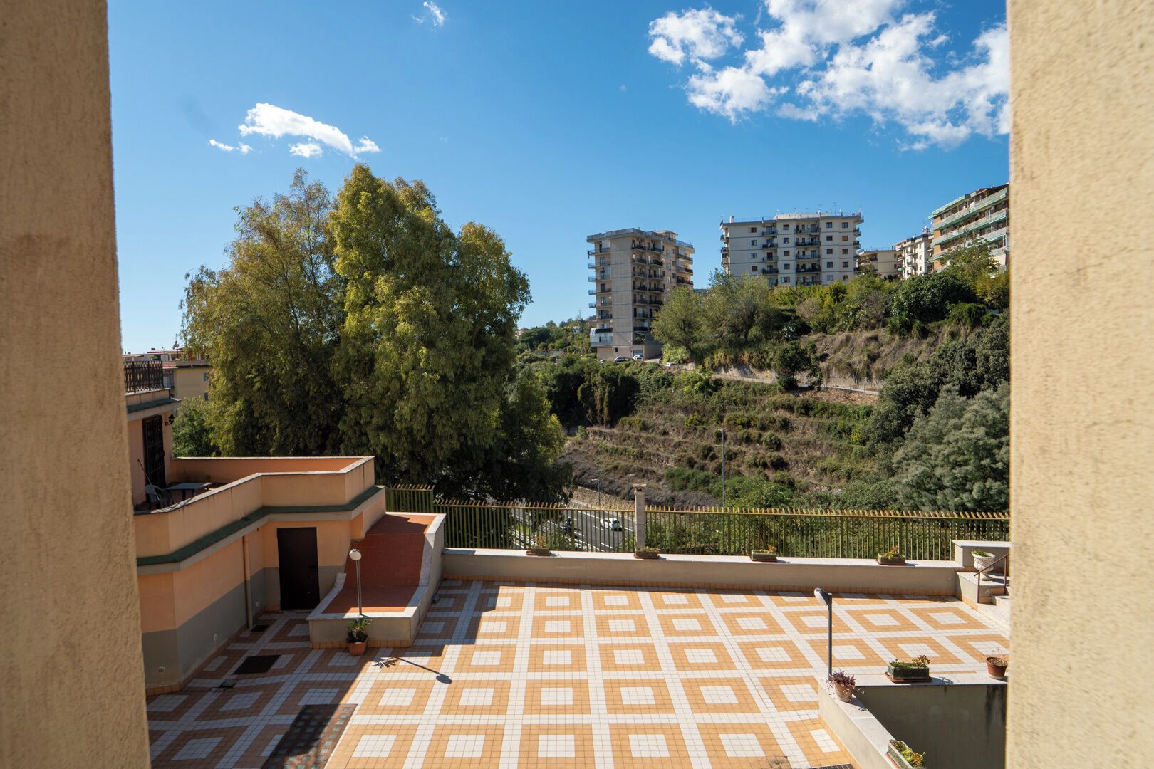 A rooftop terrace with patterned flooring, a railing, and a view of surrounding buildings and trees.