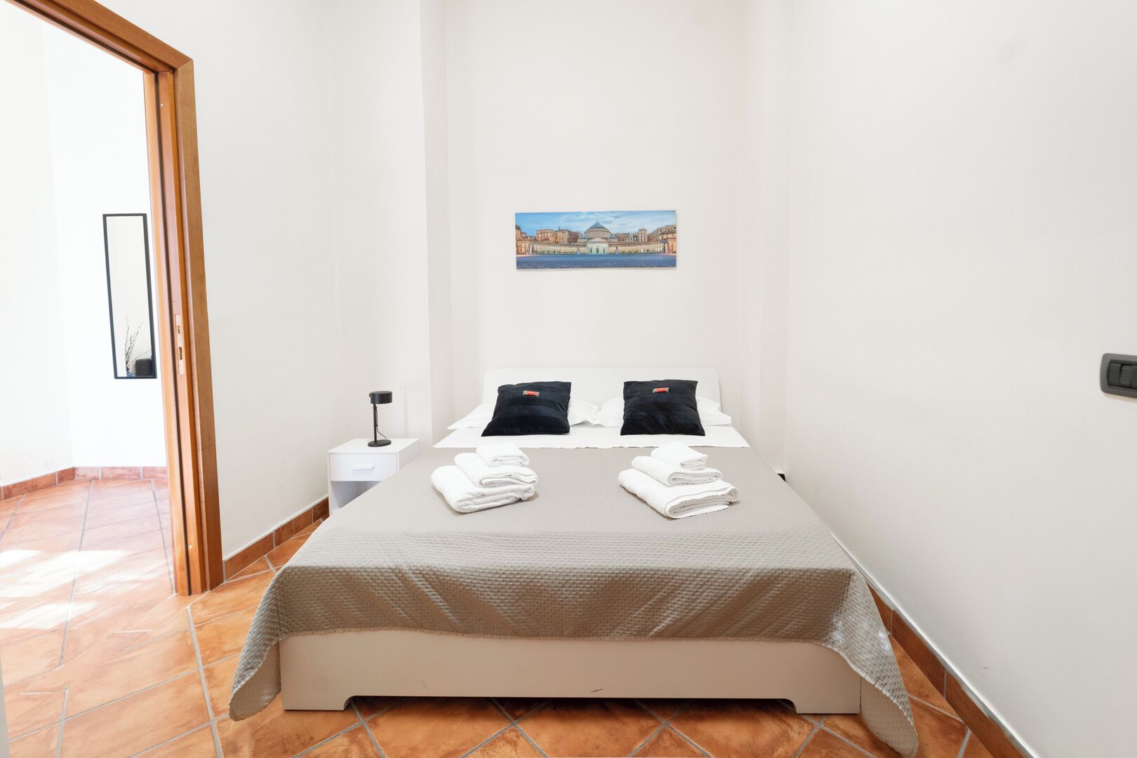 A small, clean room with a bed, two pillows, a nightstand, and a painting on the wall.