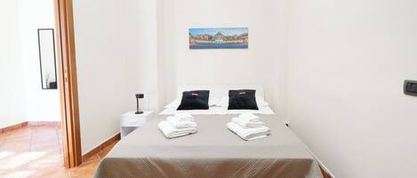A small, clean room with a bed, two pillows, a nightstand, and a painting on the wall.