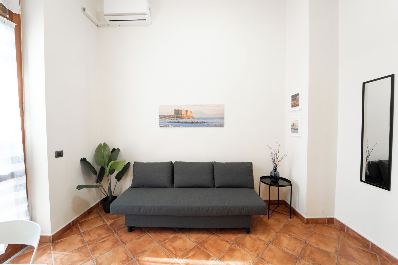 A living room with a grey sofa, a potted plant, a small round table with a vase, and a wall-mounted air conditioner.