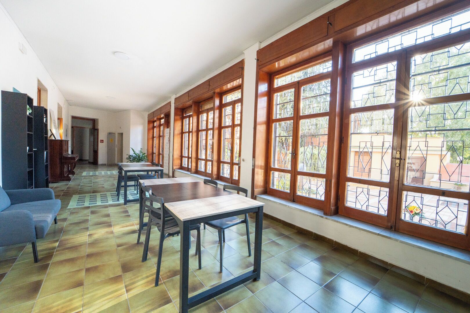 A spacious room with a dining area, large windows with wooden frames, and a tiled floor.
