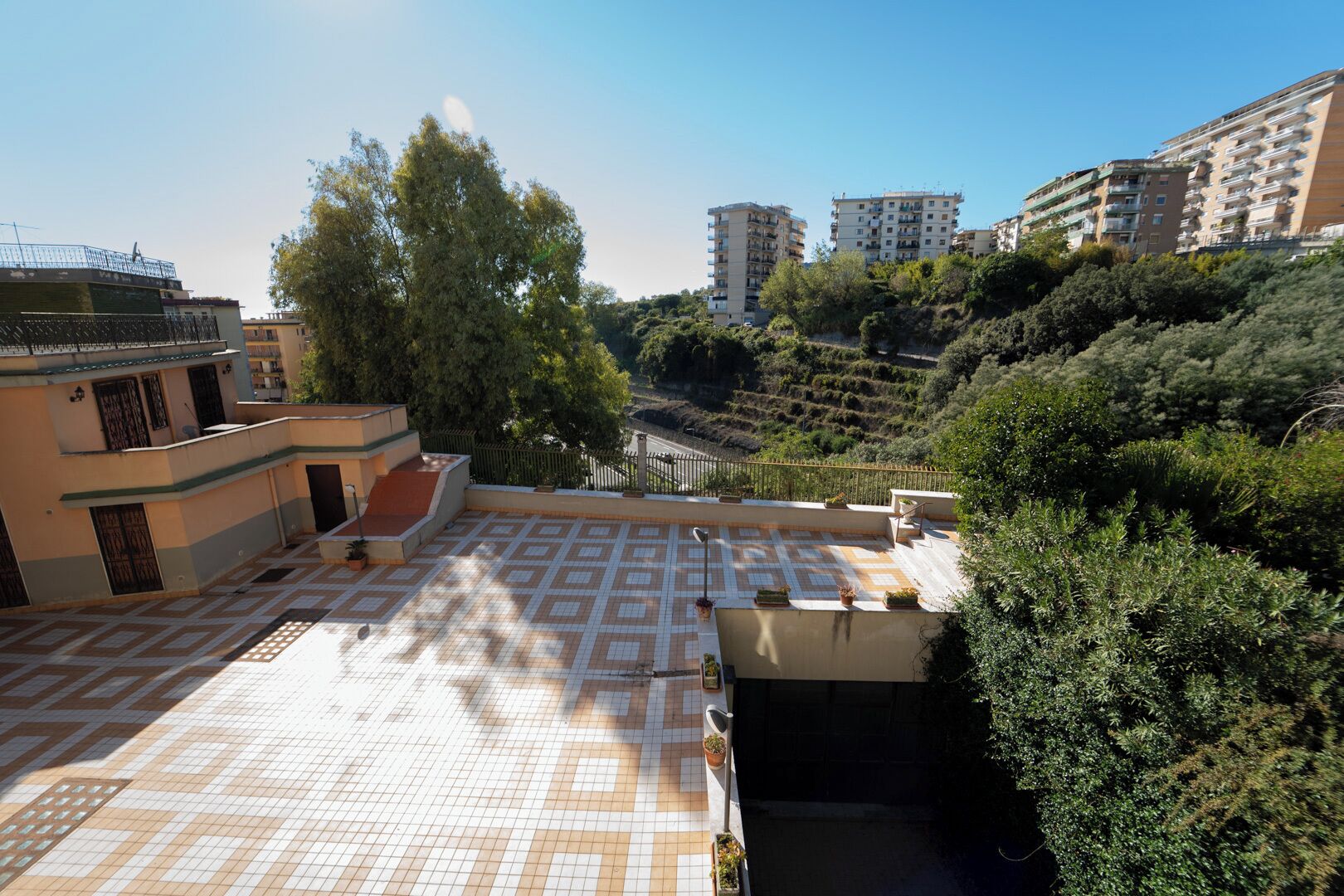 A rooftop terrace with a patterned tile floor, surrounded by greenery and residential buildings.