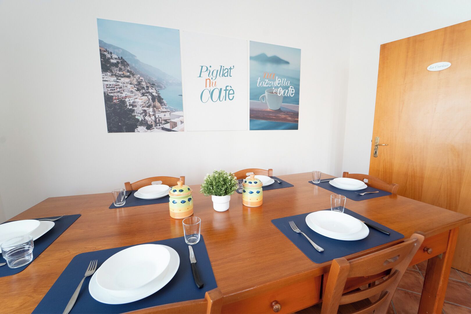 A dining area with a wooden table set for four, including plates, cutlery, and glasses. There are two wall-mounted posters, one with a coastal landscape and the other with a café scene. A small potted plant is on the table, and a wooden door is visible in the background.