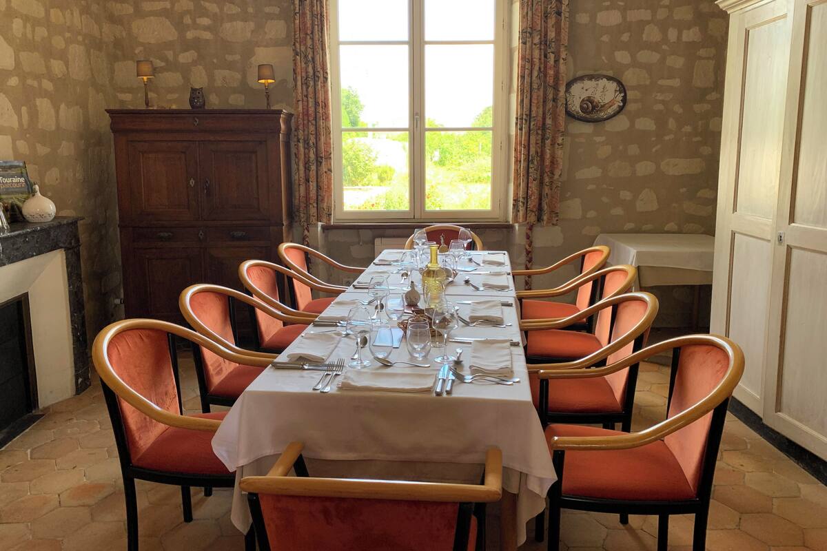 Een eetruimte met een lange tafel gedekt voor zes personen, omringd door oranje stoelen, een houten kast en een raam met gordijnen.