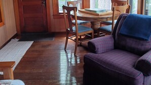 Interior d'una cabana acollidora amb parets de fusta, una taula de menjador amb cadires, un sofà morat i grans finestres que donen vista a arbres.
