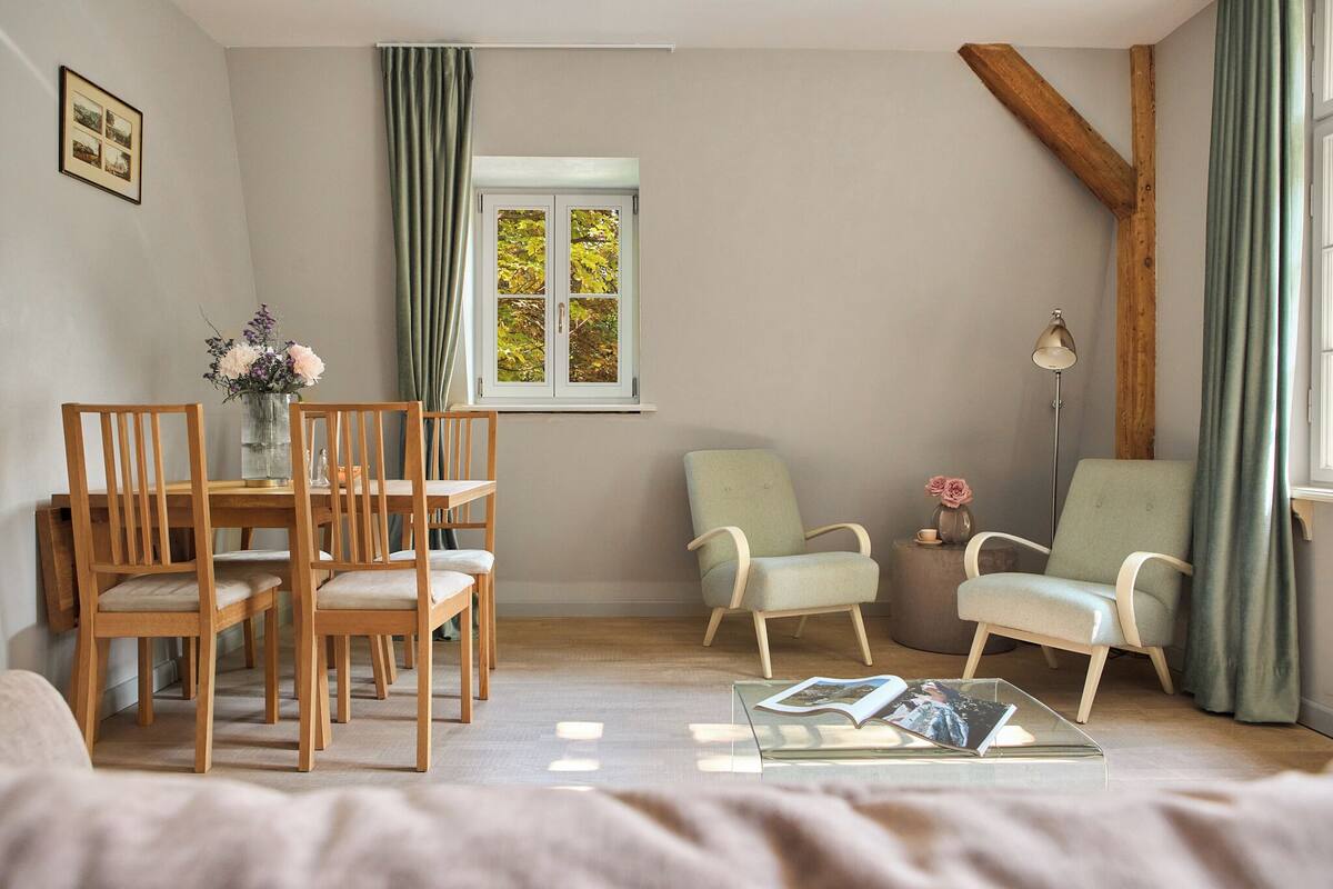 Una sala de estar acogedora con una mesa de comedor de madera, dos sillones, una mesita de noche con una lámpara y una ventana con cortinas.