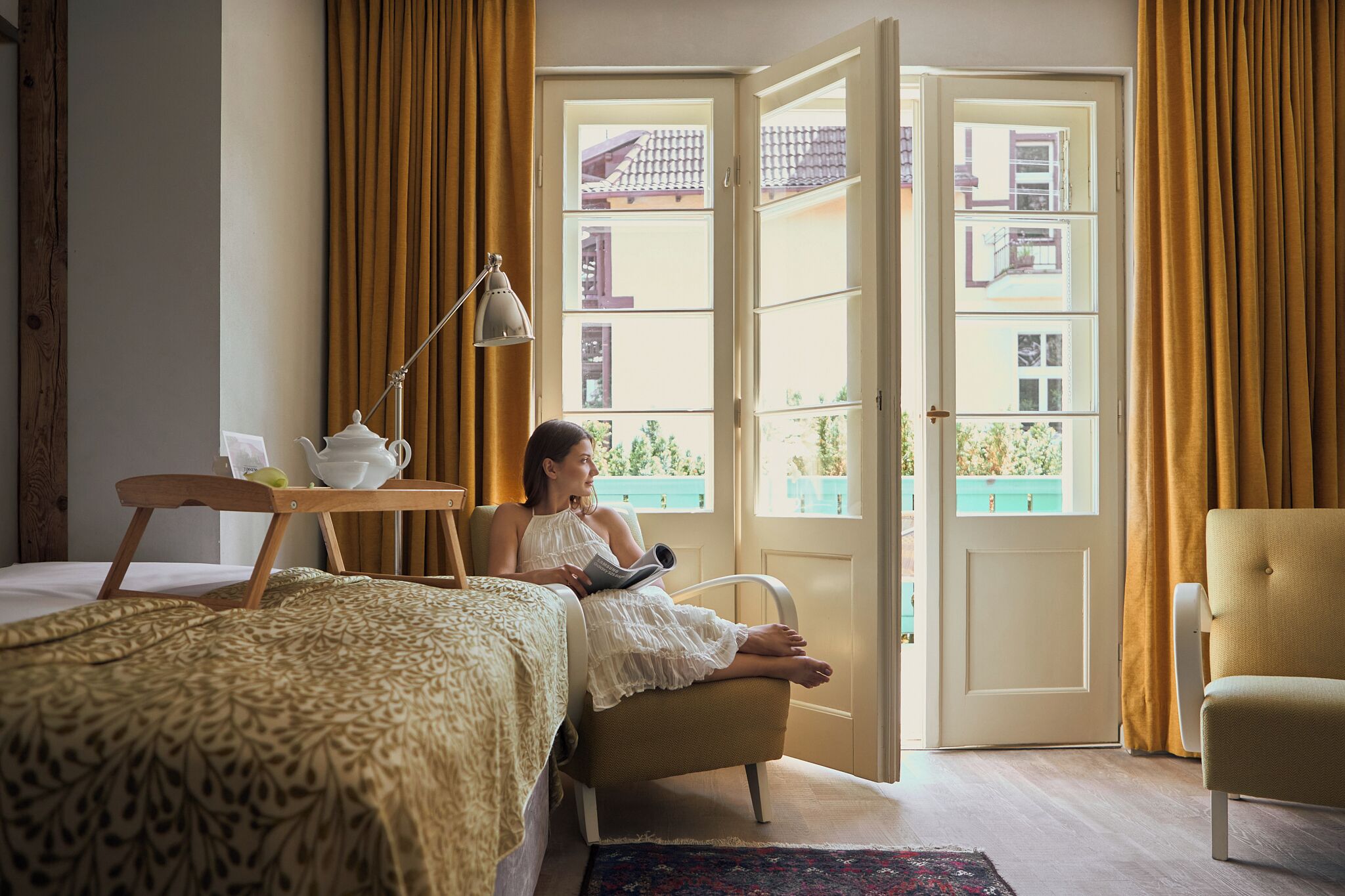 A room with a bed, a reading chair, a table with a teapot, and a window with curtains.