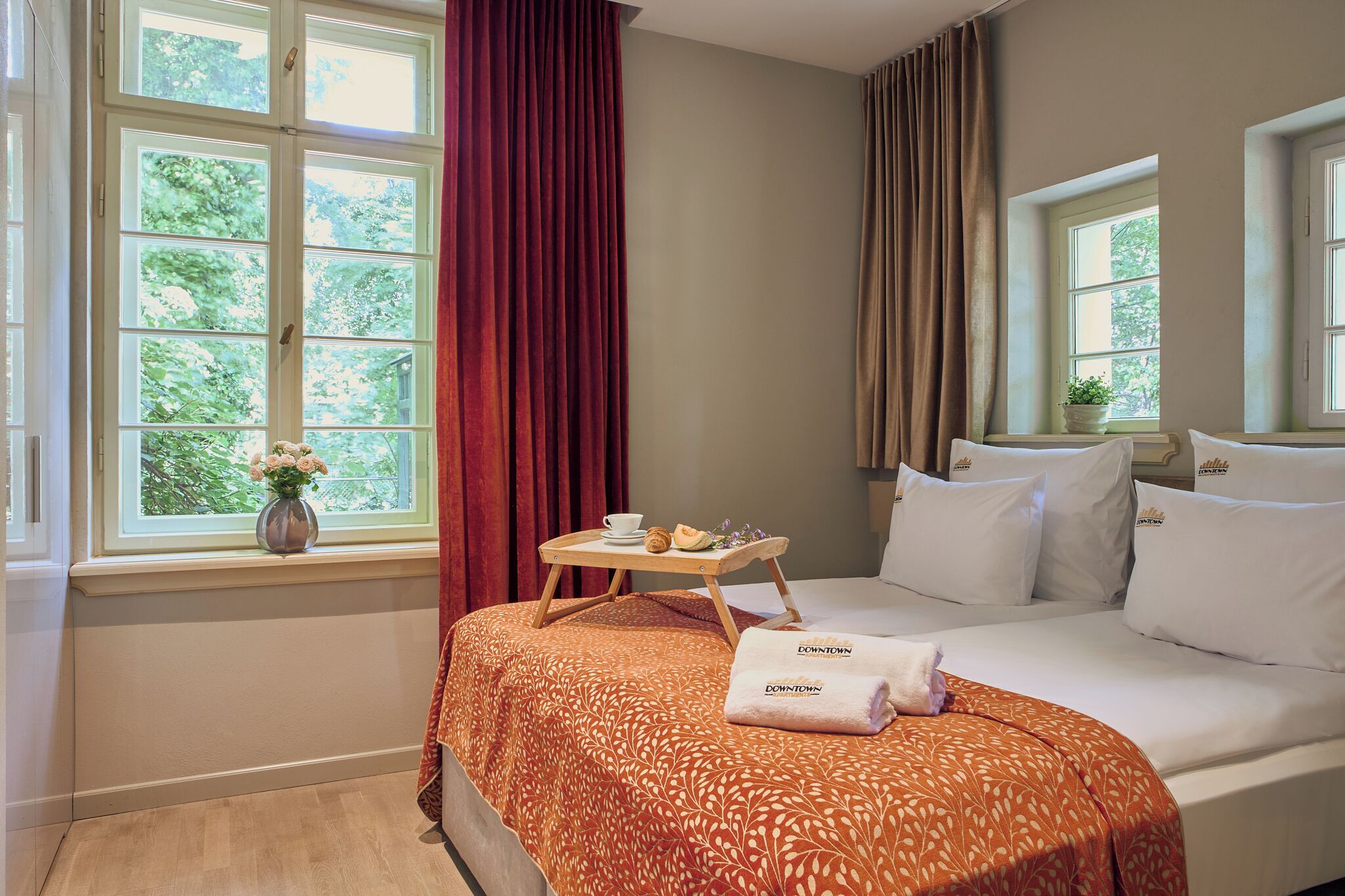 Habitación de hotel con cama, mesita de noche con bandeja de desayuno, florero con flores y ventana con cortinas.