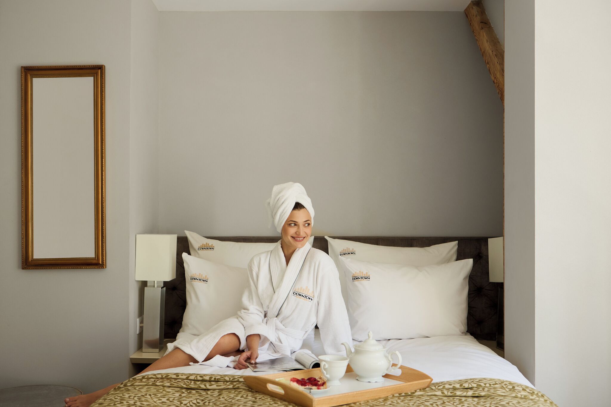 A hotel room with a bed, pillows, a bedside table with a lamp, and a tray with a teapot and a cup.