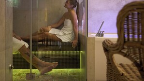 Une personne assise dans une baignoire en verre, enveloppée dans une serviette, sur un sol carrelé, avec une chaise en osier au premier plan.