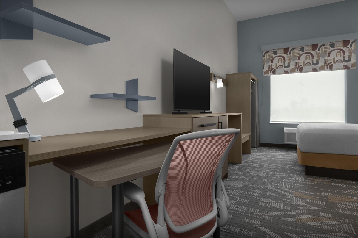 A hotel room with a desk, chair, TV, bed, and large window.