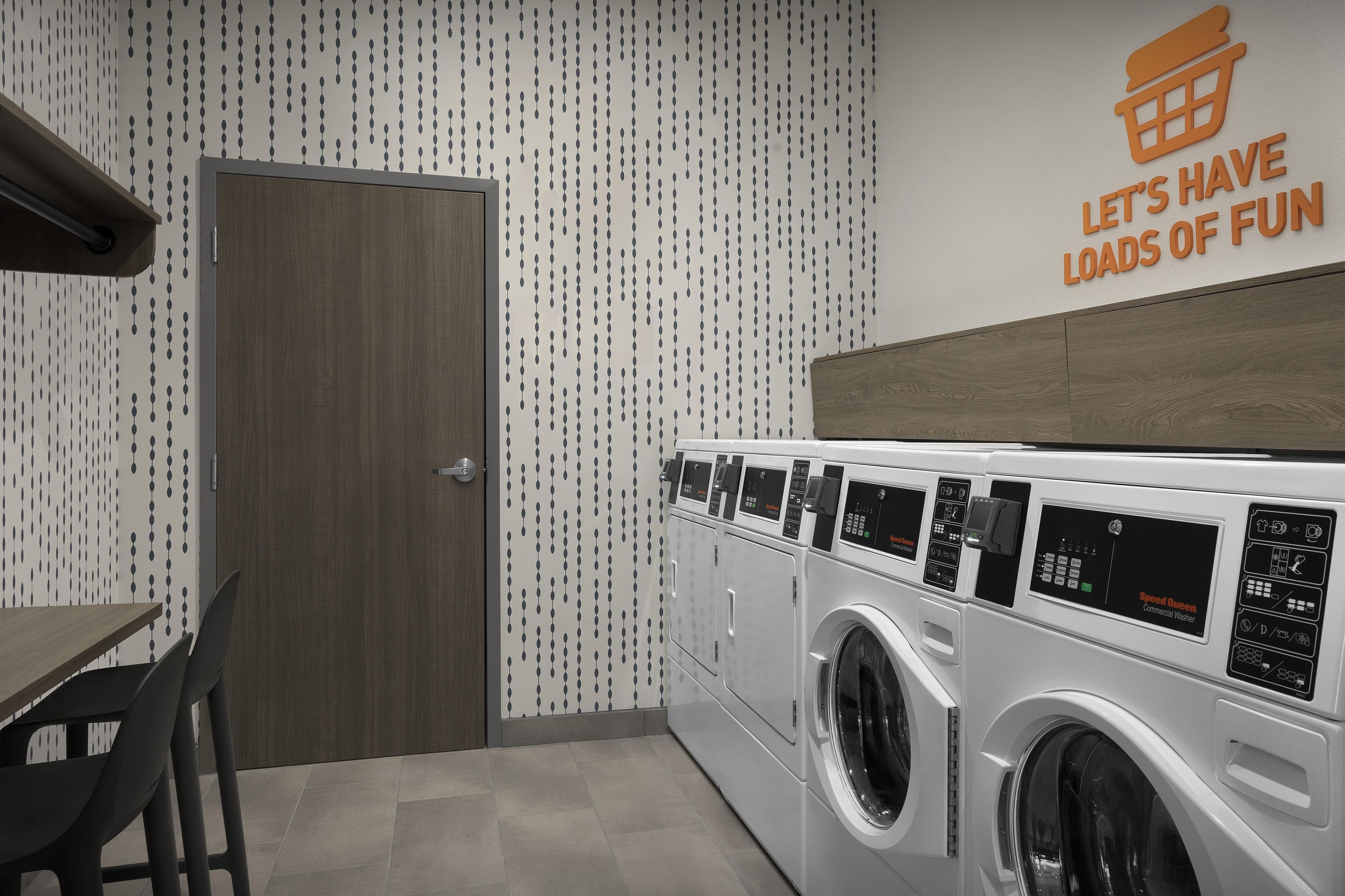 Une buanderie avec un lave-linge fixé au mur, une porte en bois et une pancarte au mur sur laquelle on peut lire : « AMUSONS-NOUS À FOND ».