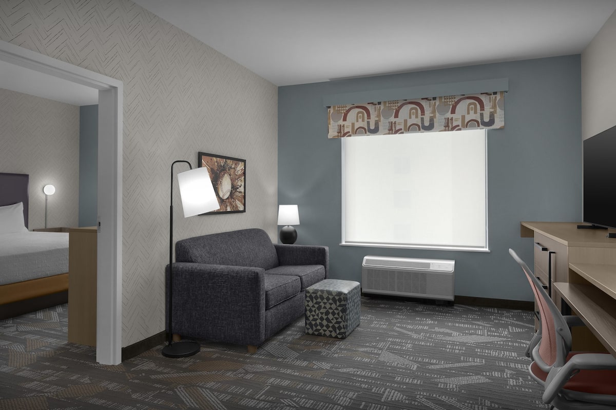 A hotel room with a bed, a sofa, a desk, a chair, a lamp, a picture on the wall, and a large window with a curtain.