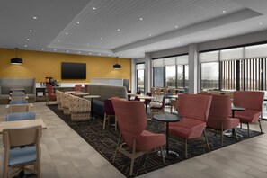 A modern indoor seating area with multiple tables and chairs, a large window offering a view, and a flat-screen TV mounted on the wall. - Home2 Suites By Hilton Taylor (Taylor)
