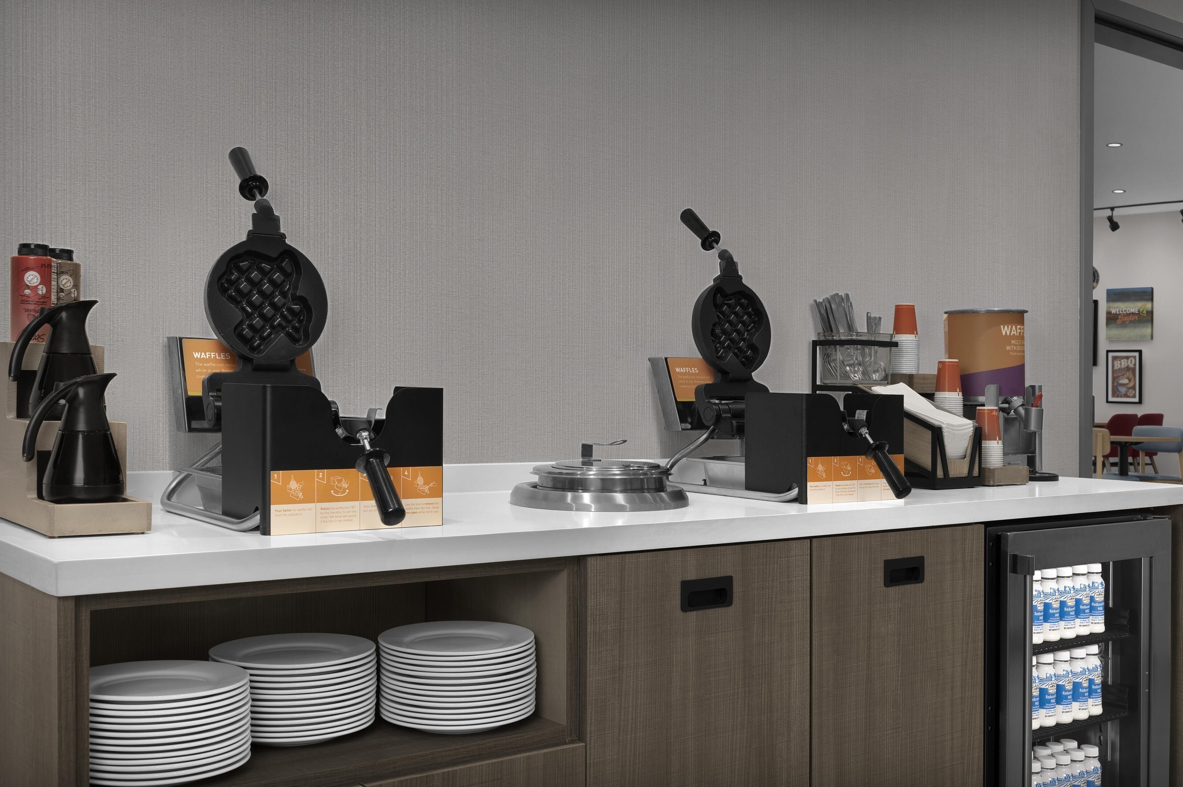 Angolo cottura con piastre per waffle, macchine da caffè e frigorifero fornito di bevande.
