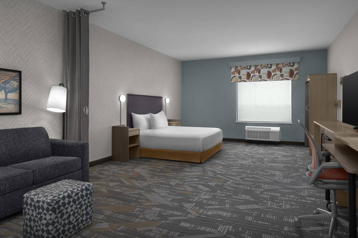 A hotel room with a bed, a desk, a chair, a sofa, a patterned carpet, a window with curtains, and a wall-mounted TV.