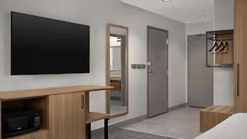 A modern hotel room with a flat-screen TV, a wooden cabinet with a microwave, a mirror, a wardrobe, and a small wooden stool.