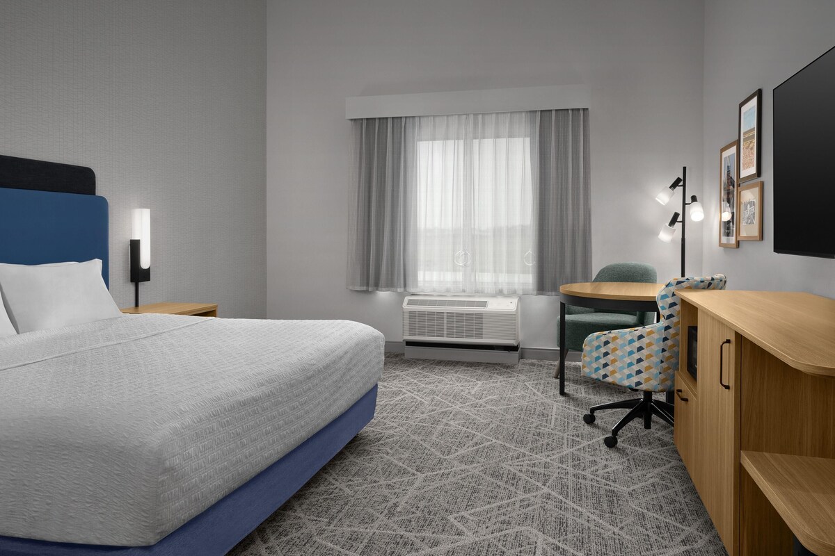 A hotel room with a bed, a desk, a chair, a window with curtains, a wall-mounted TV, and a radiator.