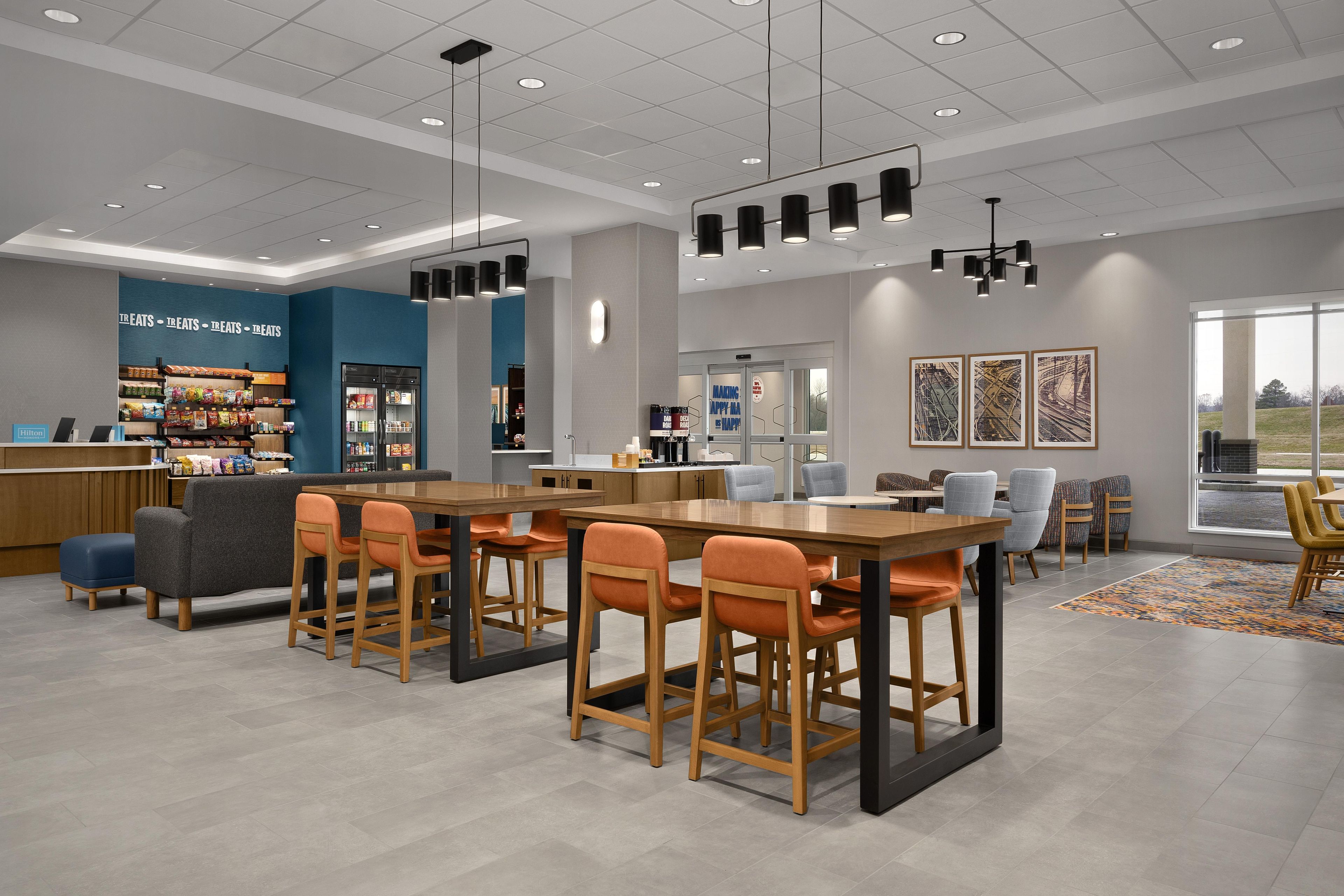 A modern hotel lobby with a central dining area, a minibar, and a view of the outdoors.