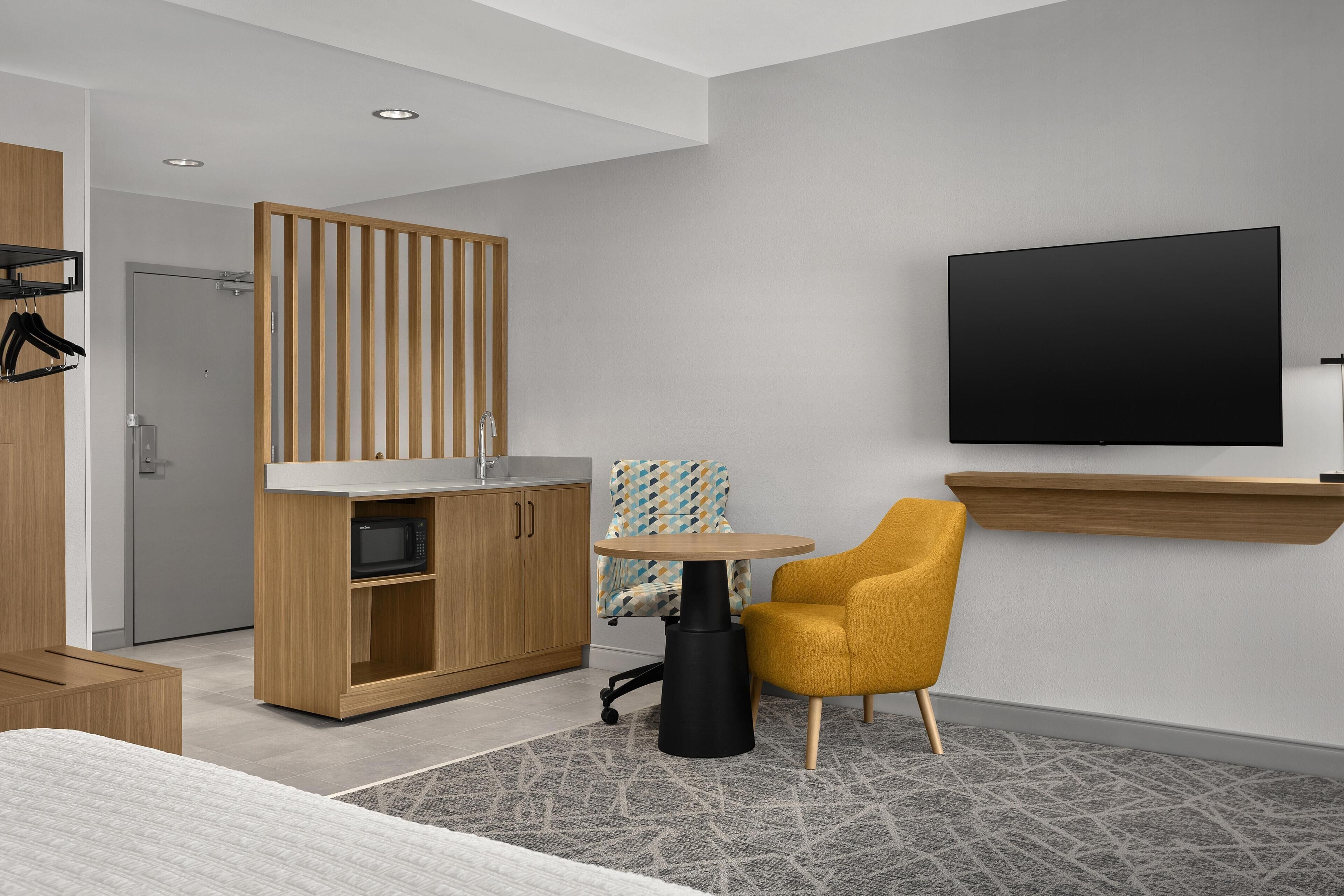 Habitación de hotel moderna con cama, armario de madera, kitchenette, mesa redonda con una silla y televisor montado.