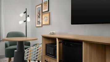 Habitación de hotel compacta con escritorio de madera, microondas, refrigerador negro, sillón verde, mesita con cojín estampado y televisor montado en la pared.