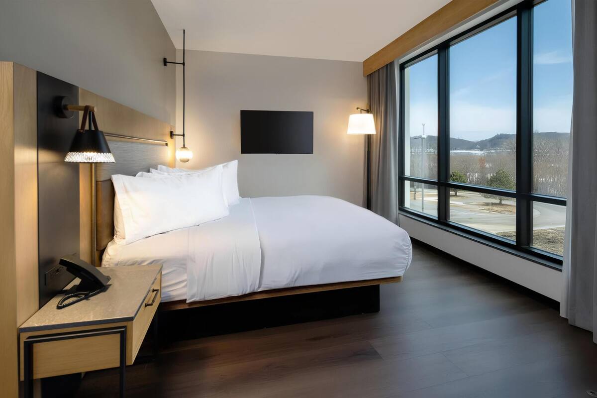 Une chambre d’hôtel moderne avec un grand lit, une table de chevet, une lampe et une vue par la fenêtre.
