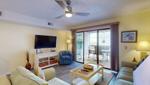 A living room with a flat-screen TV, a ceiling fan, a sofa, a coffee table, and a sliding glass door leading to an outdoor area.