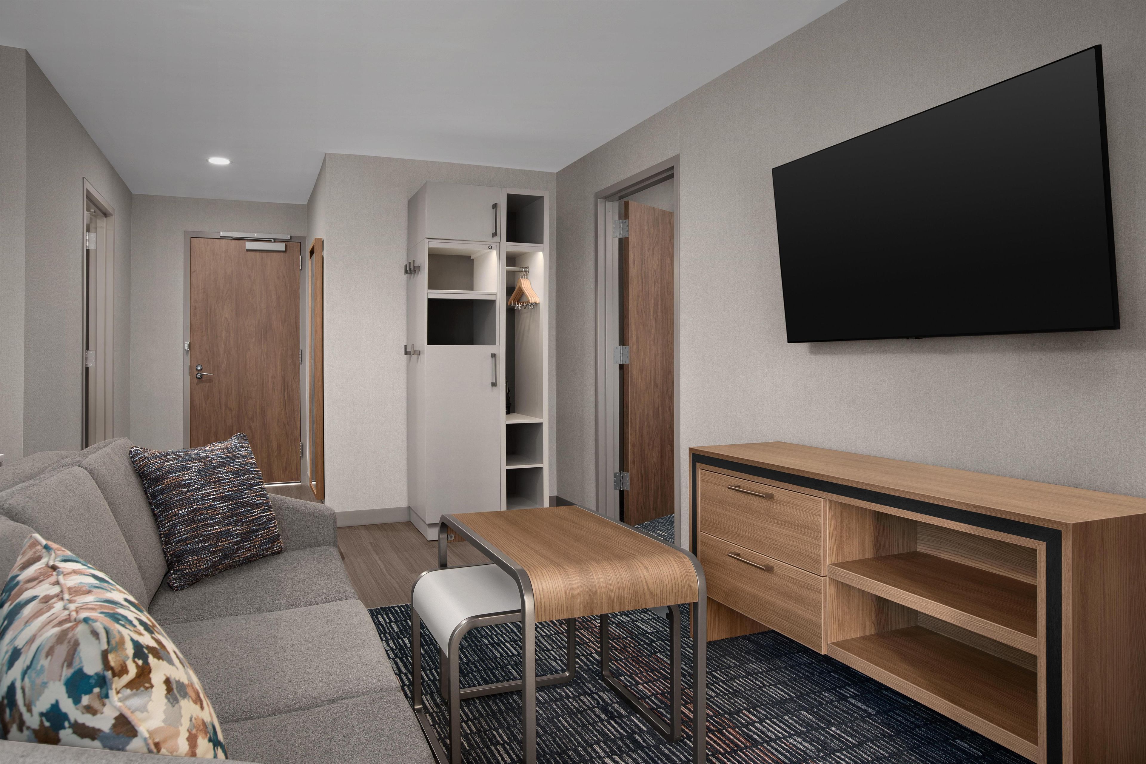 Een moderne hotelkamer met een grijze bank, een klein houten tafeltje, een aan de muur bevestigde televisie en een garderobe met spiegel.