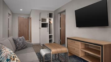Een moderne hotelkamer met een grijze bank, een klein houten tafeltje, een aan de muur bevestigde televisie en een garderobe met spiegel.