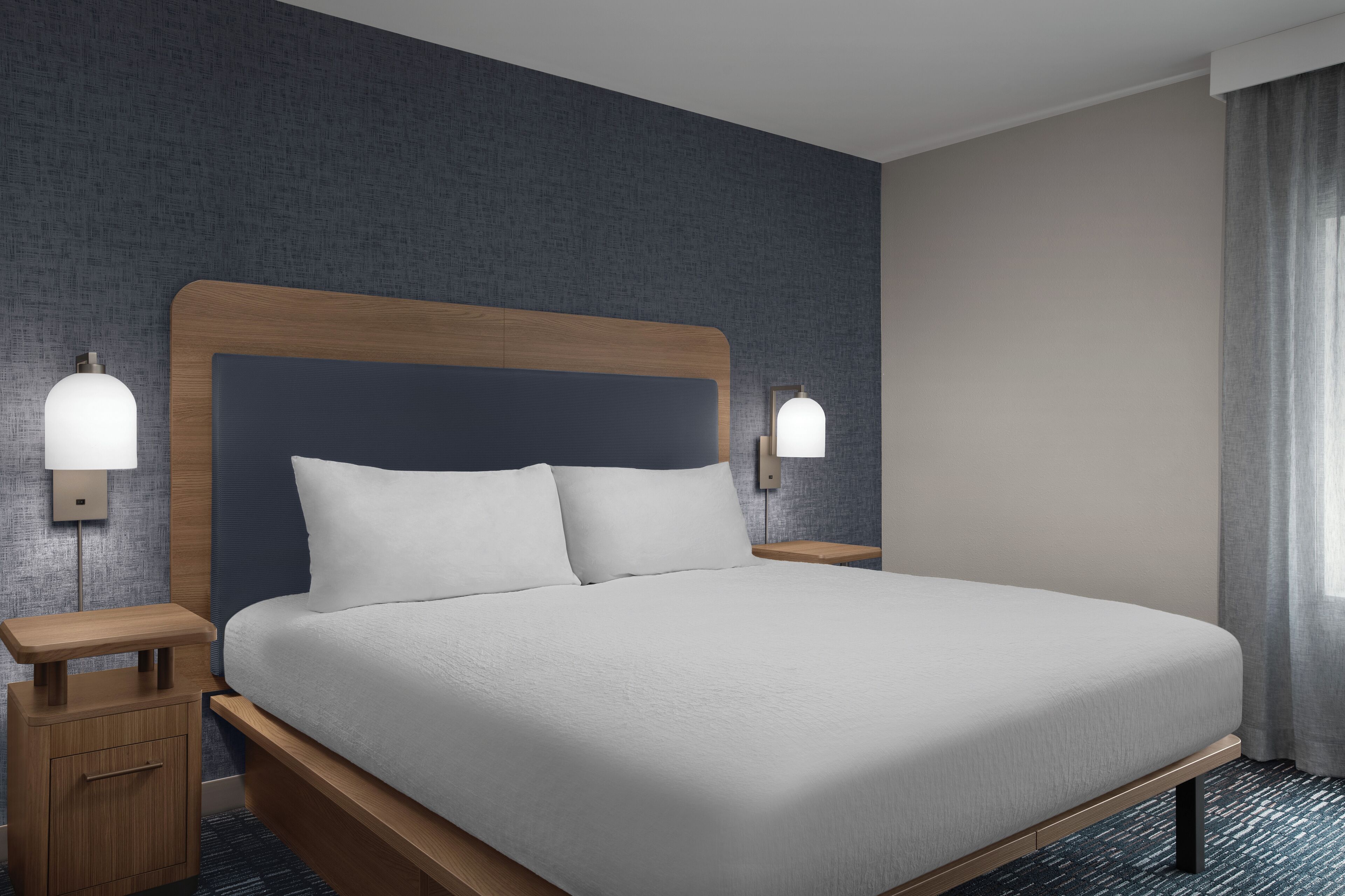 A modern hotel room with a large bed, wooden headboard, two bedside tables with lamps, and a textured wall.