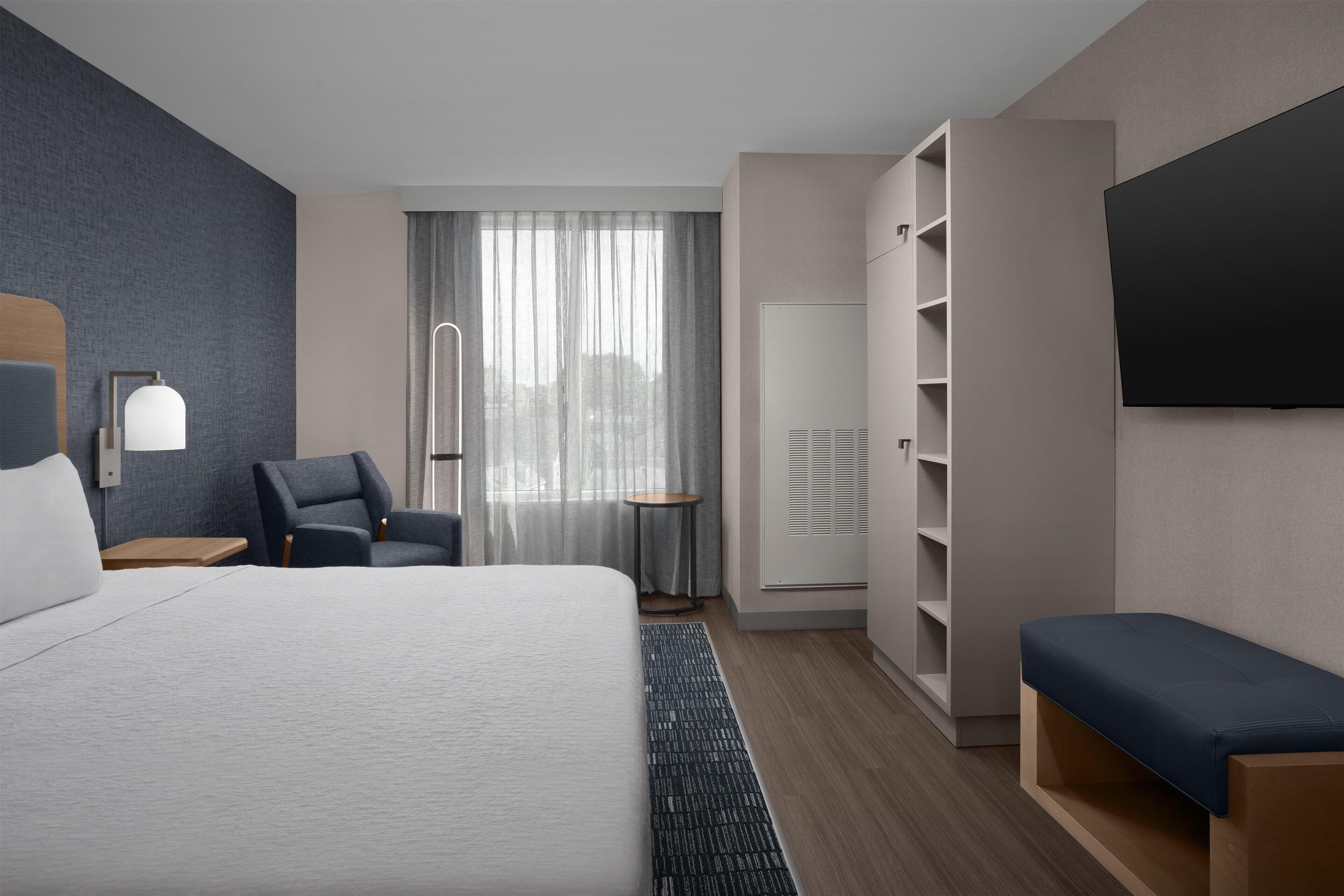 A modern hotel room with a large bed, a seating area, a wardrobe, a television, and a window with curtains.