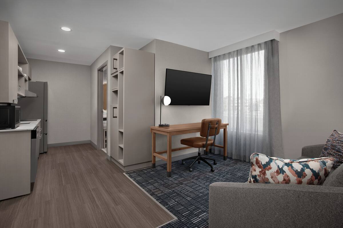A modern hotel room with a kitchenette, a flat-screen TV mounted on the wall, a wooden desk, a comfortable sofa, and a large window with sheer curtains.