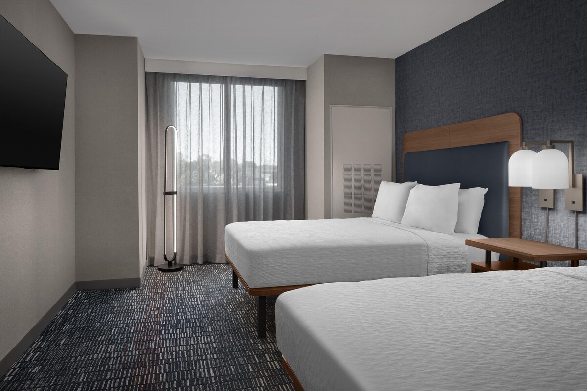 Habitación de hotel con dos camas, un escritorio, una silla, un televisor montado en la pared y una ventana con cortinas.