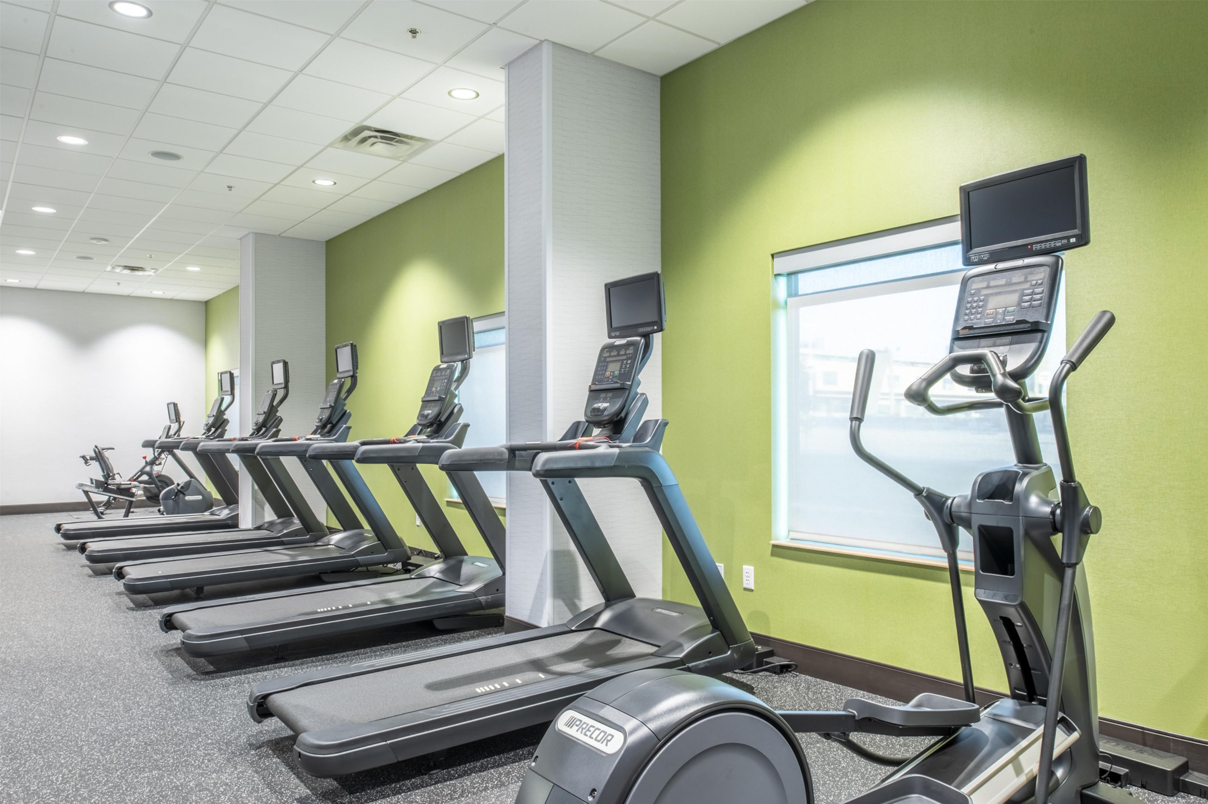 A fitness center with multiple treadmills and elliptical machines, a television mounted on the wall, and a window providing natural light.